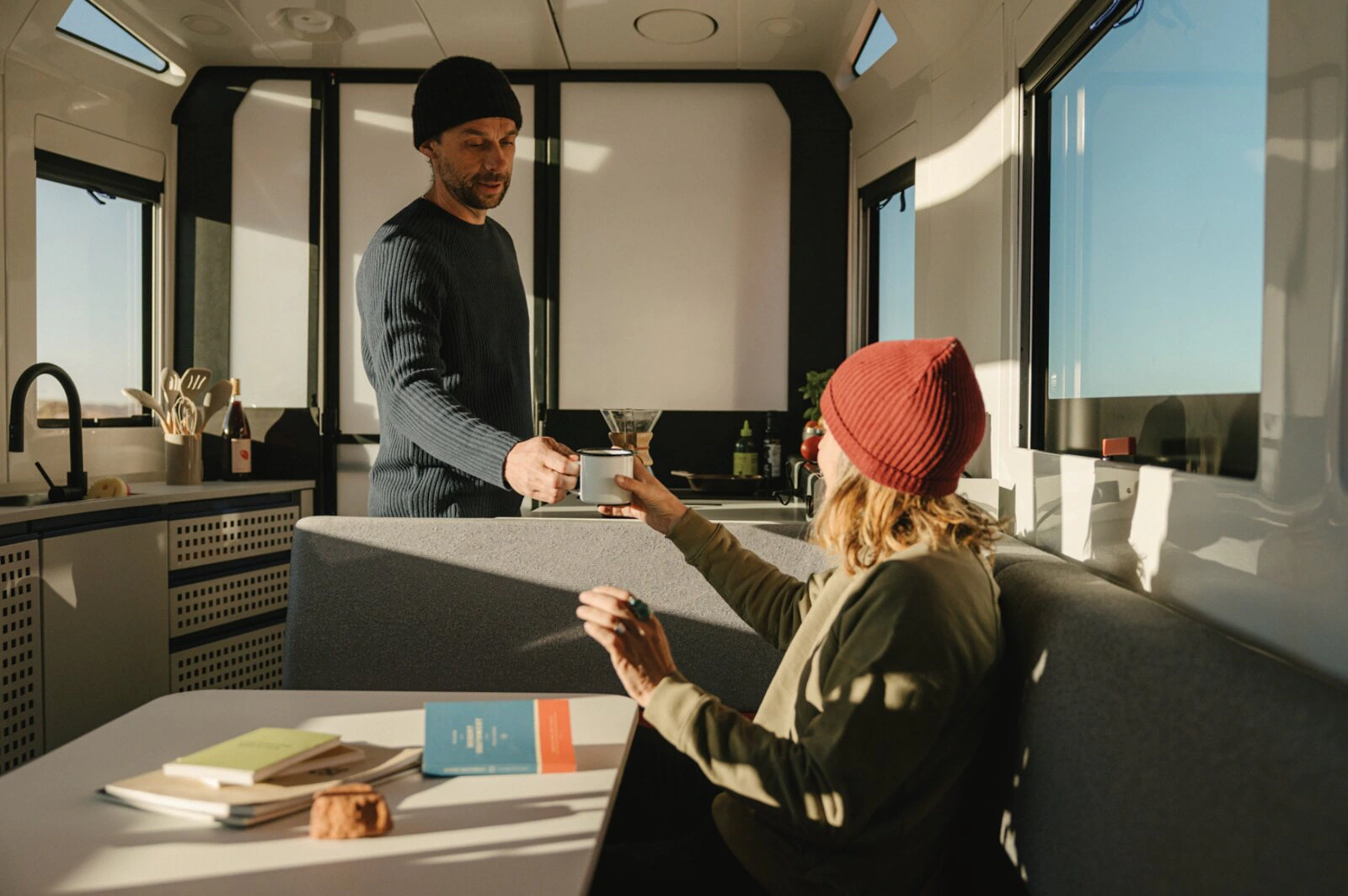 Two people sharing coffee inside the Lightship trailer in warm morning light.