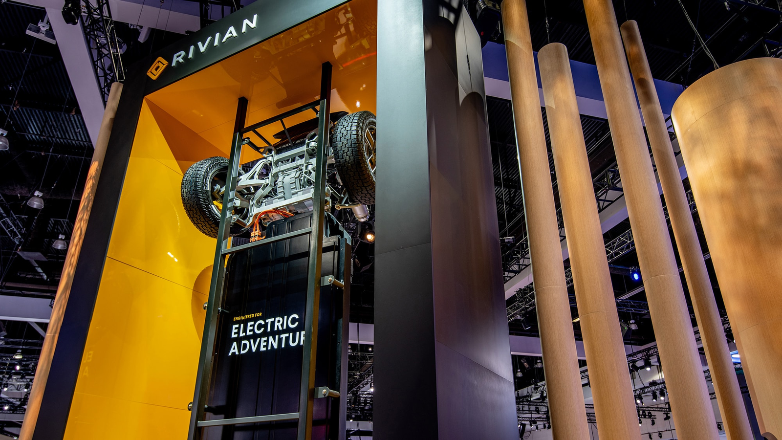 A Rivian electric skateboard chassis displayed vertically between timber columns at the auto show.