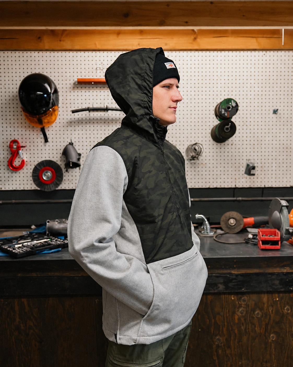 A model wearing the Terrascout gray wool and dark camouflage-panel anorak, with its hood over a black knit hat, stands in profile with his hands in the front pocket against a workshop pegboard.