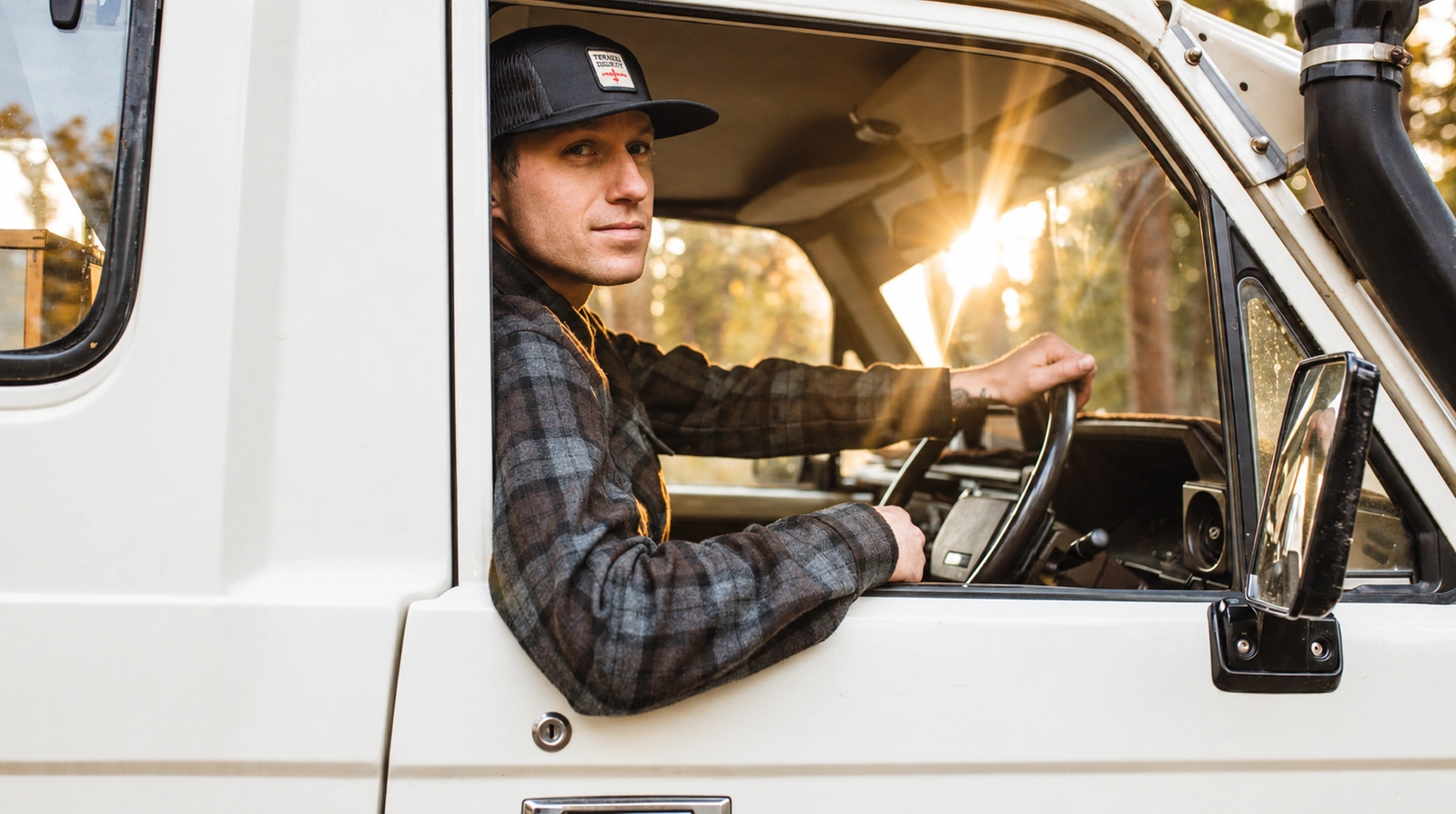 A model wearing a gray plaid Terrascout shirtjacket and branded cap at the wheel of a white vintage Land Cruiser, with warm sunlight through the windshield.