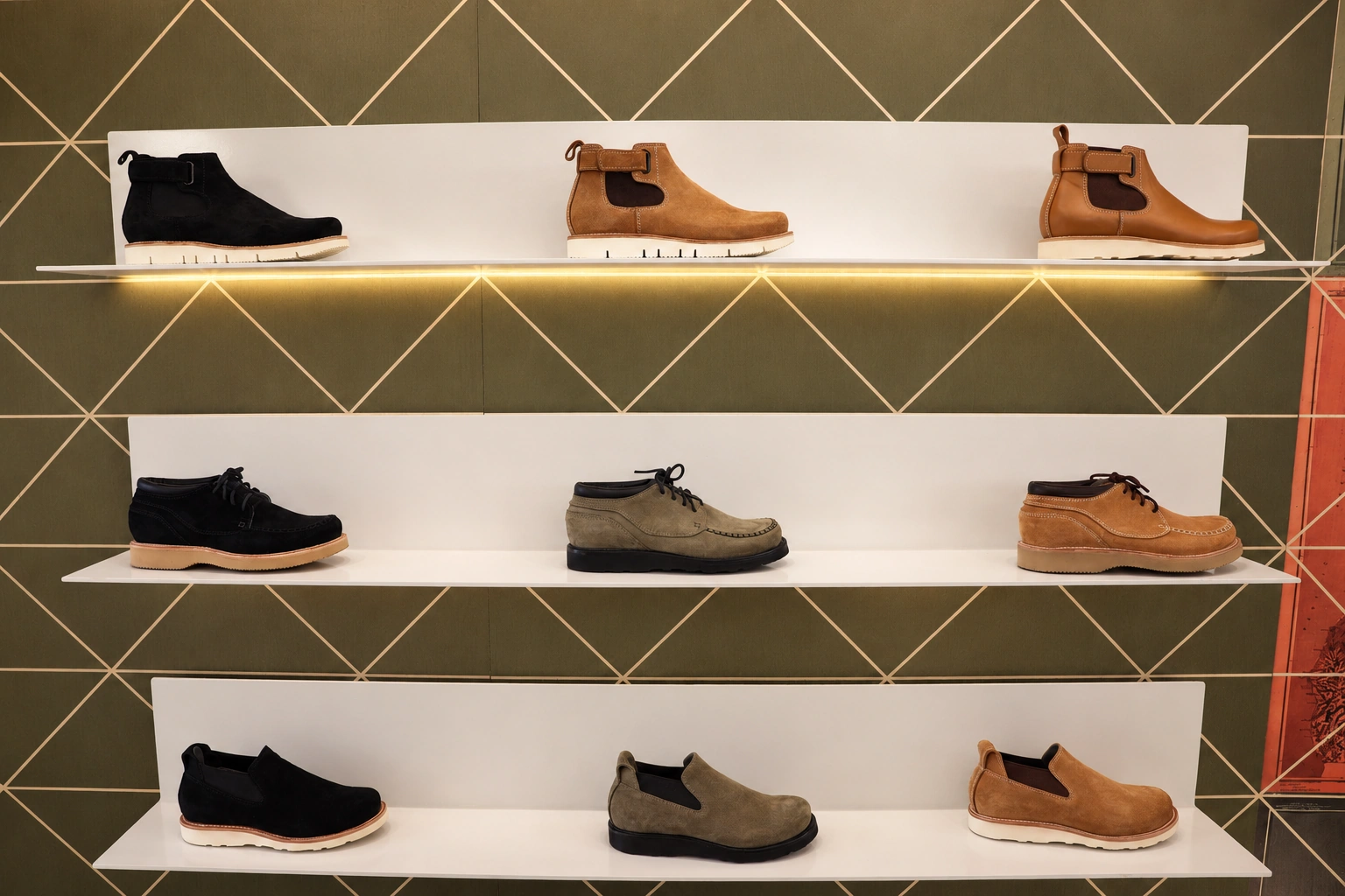 Nine Terrascout footwear designs in black, olive and tan leather and suede, arranged across three white shelves against the geometric olive wall.