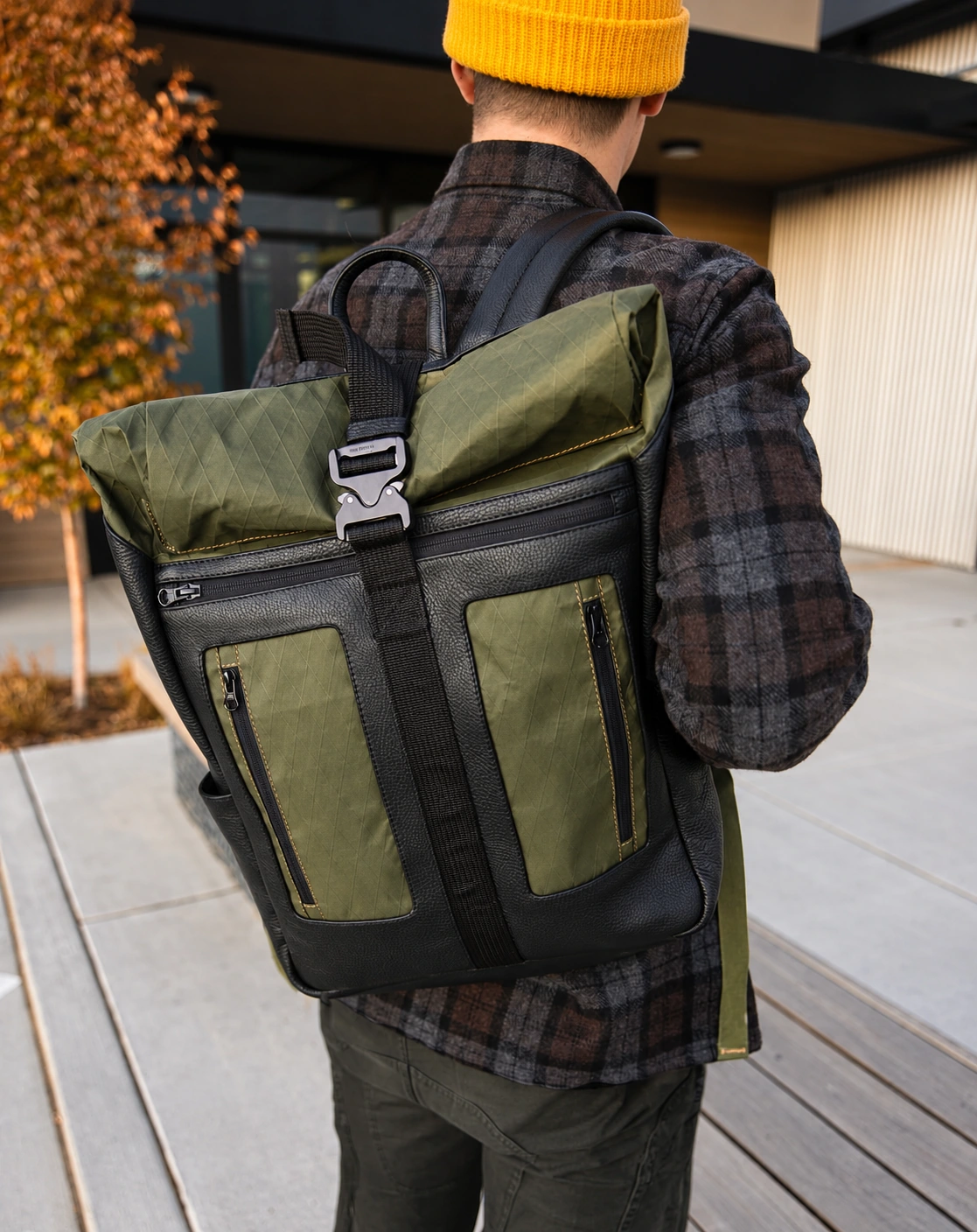 Close view of a black and olive Terrascout rolltop backpack worn over a dark plaid shirtjacket, showing the metal buckle, front zip pockets, pebbled surfaces and diamond-pattern technical fabric beneath a mustard knit hat.