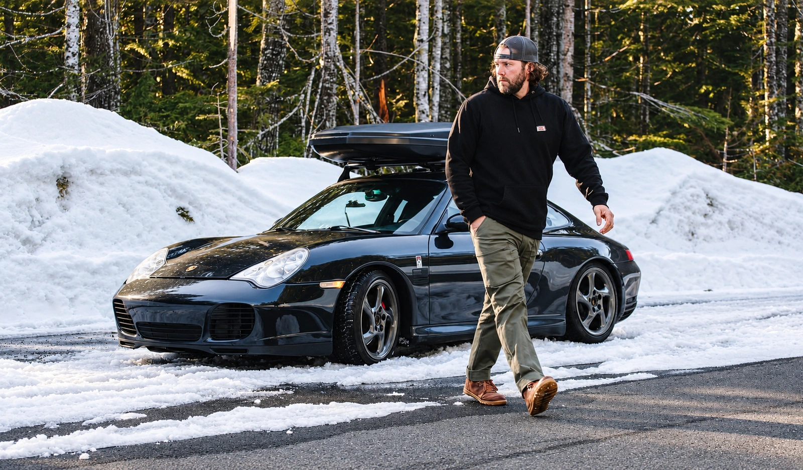A man in a black Terrascout hoodie, olive utility trousers and brown shoes walks along a snowy forest road in front of a dark Porsche with a roof box.