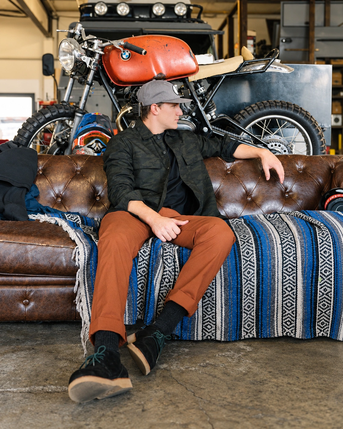 A model in a dark patterned Terrascout overshirt, rust trousers and dark wedge-soled shoes sits on a brown leather couch draped with a blue striped blanket, with an orange motorcycle in the workshop behind him.